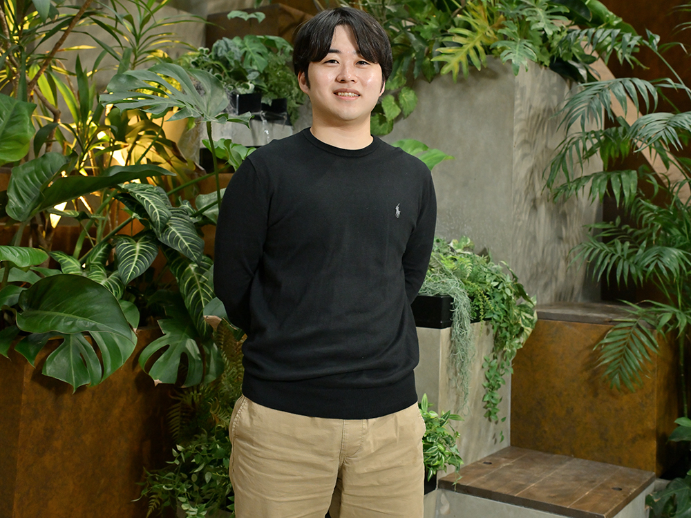 A man in casual attire stands in an indoor garden environment surrounded by lush green plants.
