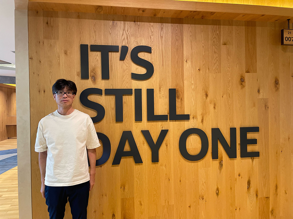 A man stands in front of a wooden wall with large black letters spelling out 'IT'S STILL DAY ONE' at an AWS office.