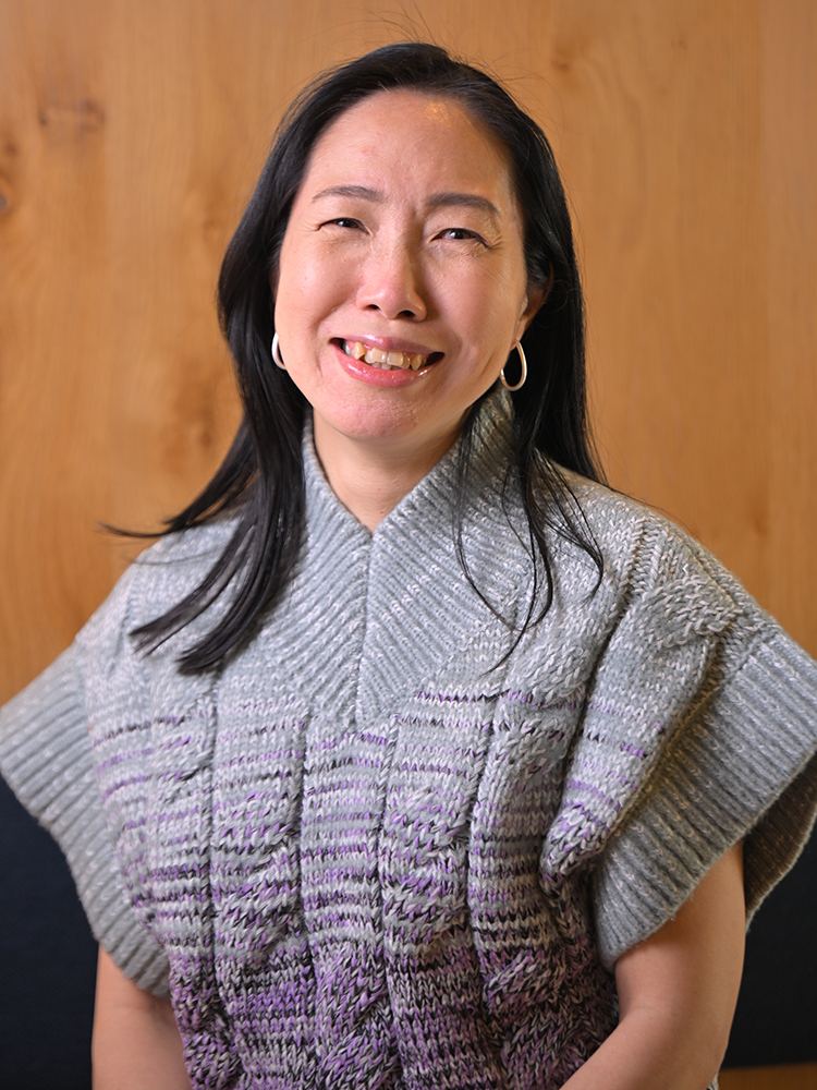 A woman smiles while wearing a gray and purple knit sweater, with a wood background behind her.