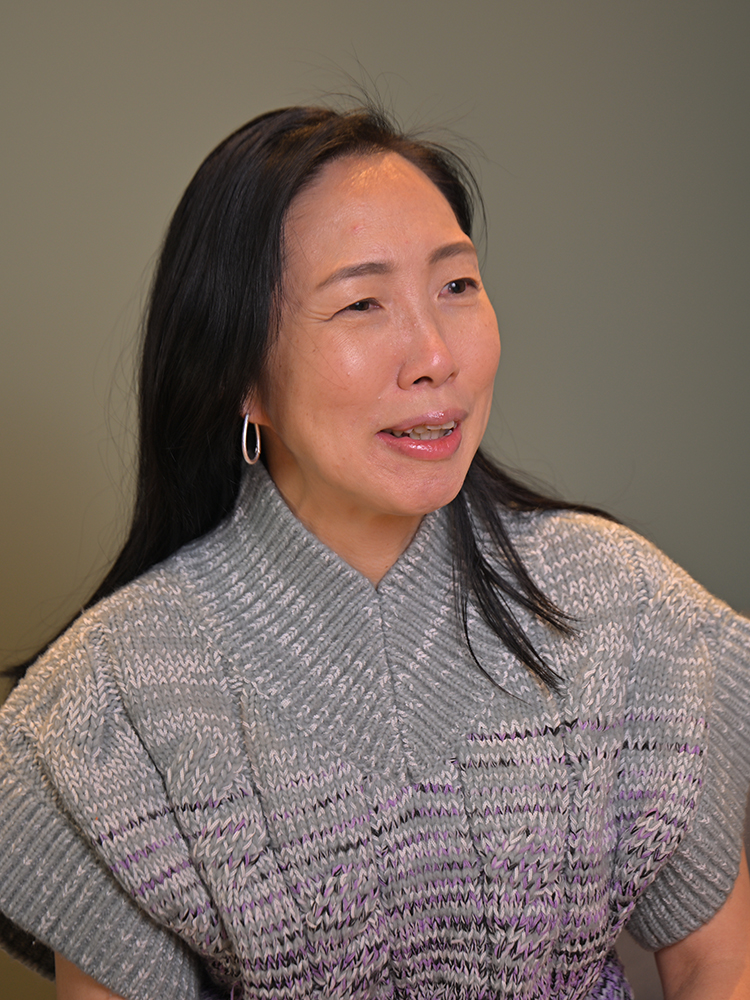 A woman with long dark hair wearing a gray knit sweater, photographed in front of a plain background.