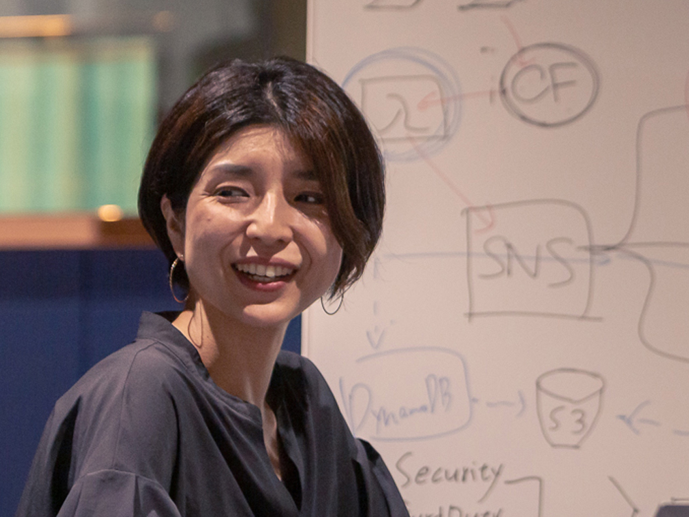 A woman presenting and smiling in front of a whiteboard covered with technical diagrams and notes.