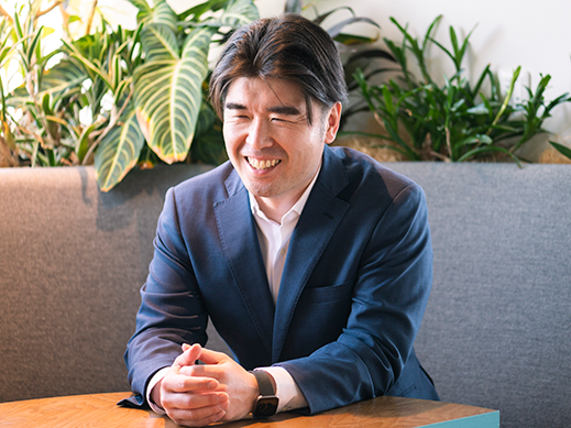 A smiling man in a blue suit sitting at a desk with several indoor plants in the background.