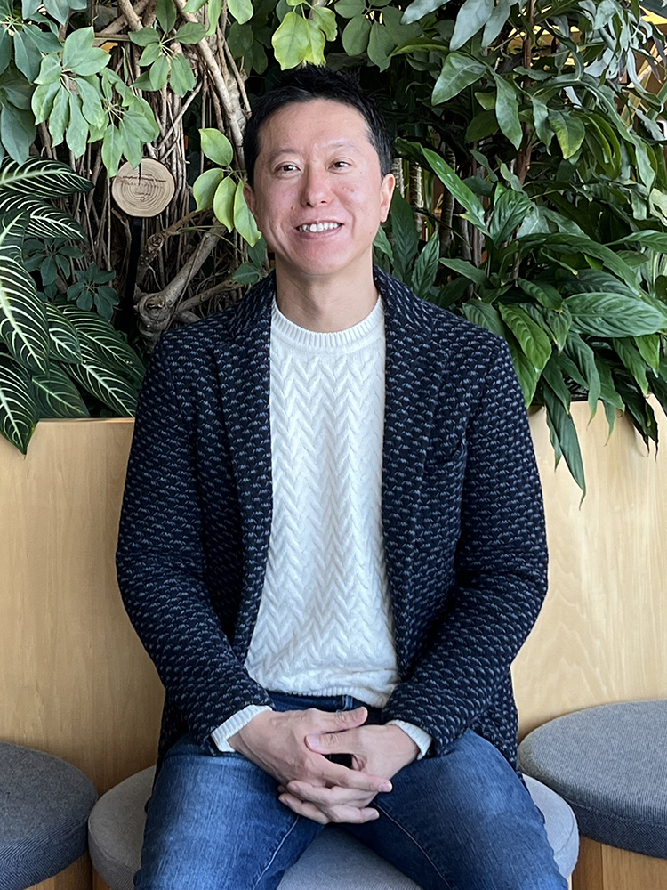 A man is sitting indoors in casual attire with a background of lush green plants. He is smiling, wearing a white sweater and dark jacket, with blue jeans, and seated on a gray bench. The setting has a relaxed and natural atmosphere.