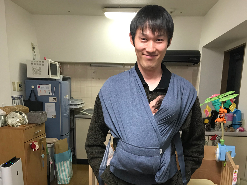 A man standing in a kitchen, smiling, while holding a baby in a carrier on his chest.