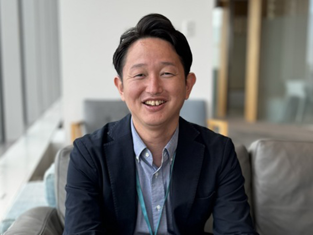 A smiling man sits in an office environment, dressed in a light shirt and casual dark blazer.