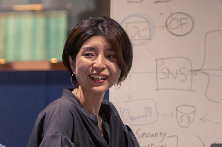 A woman smiling and participating in a discussion in front of a whiteboard with technical diagrams, likely in a workplace or office setting.