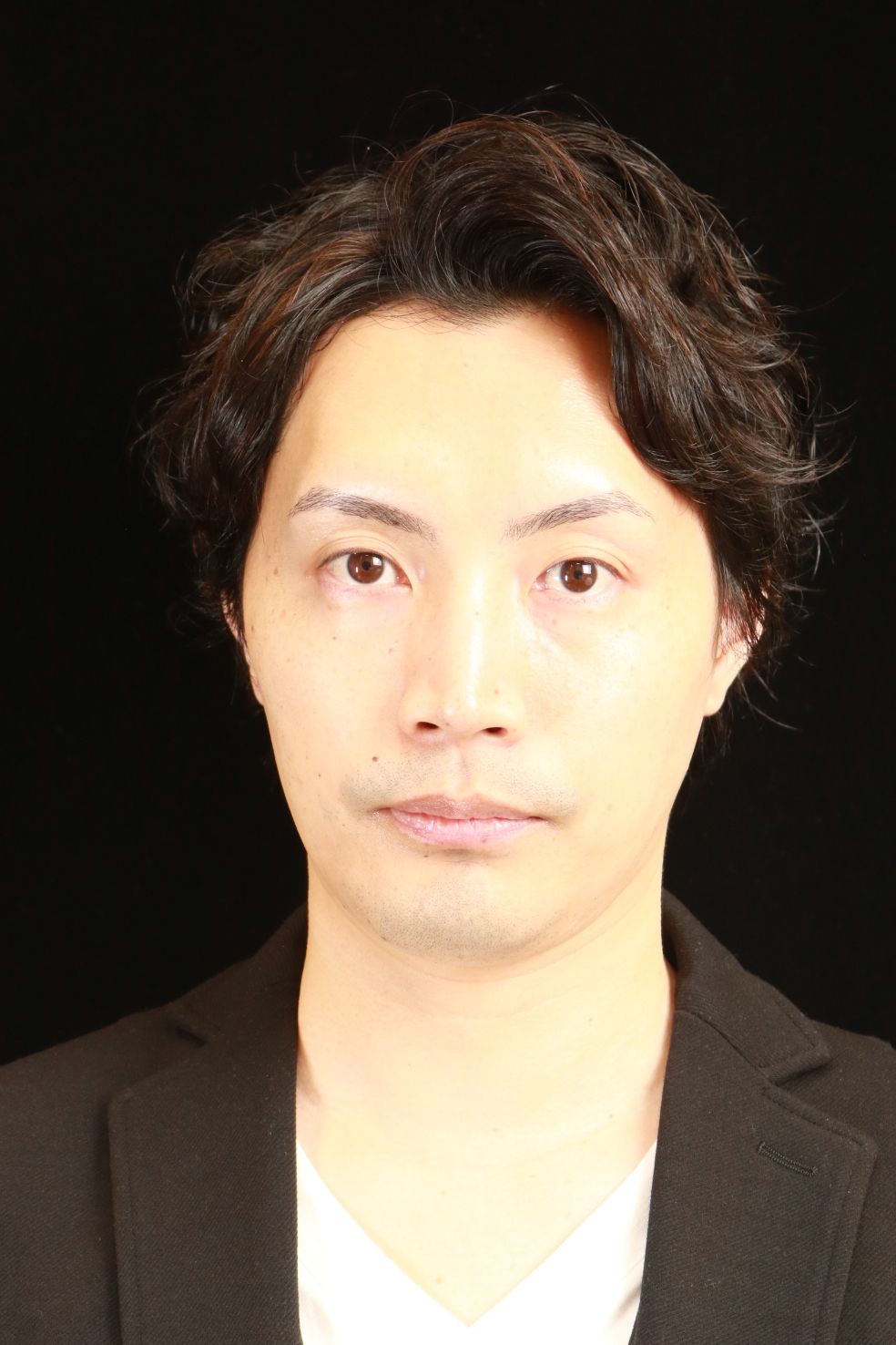 A portrait of an Asian man with short, wavy hair, wearing a black jacket, photographed against a black background.