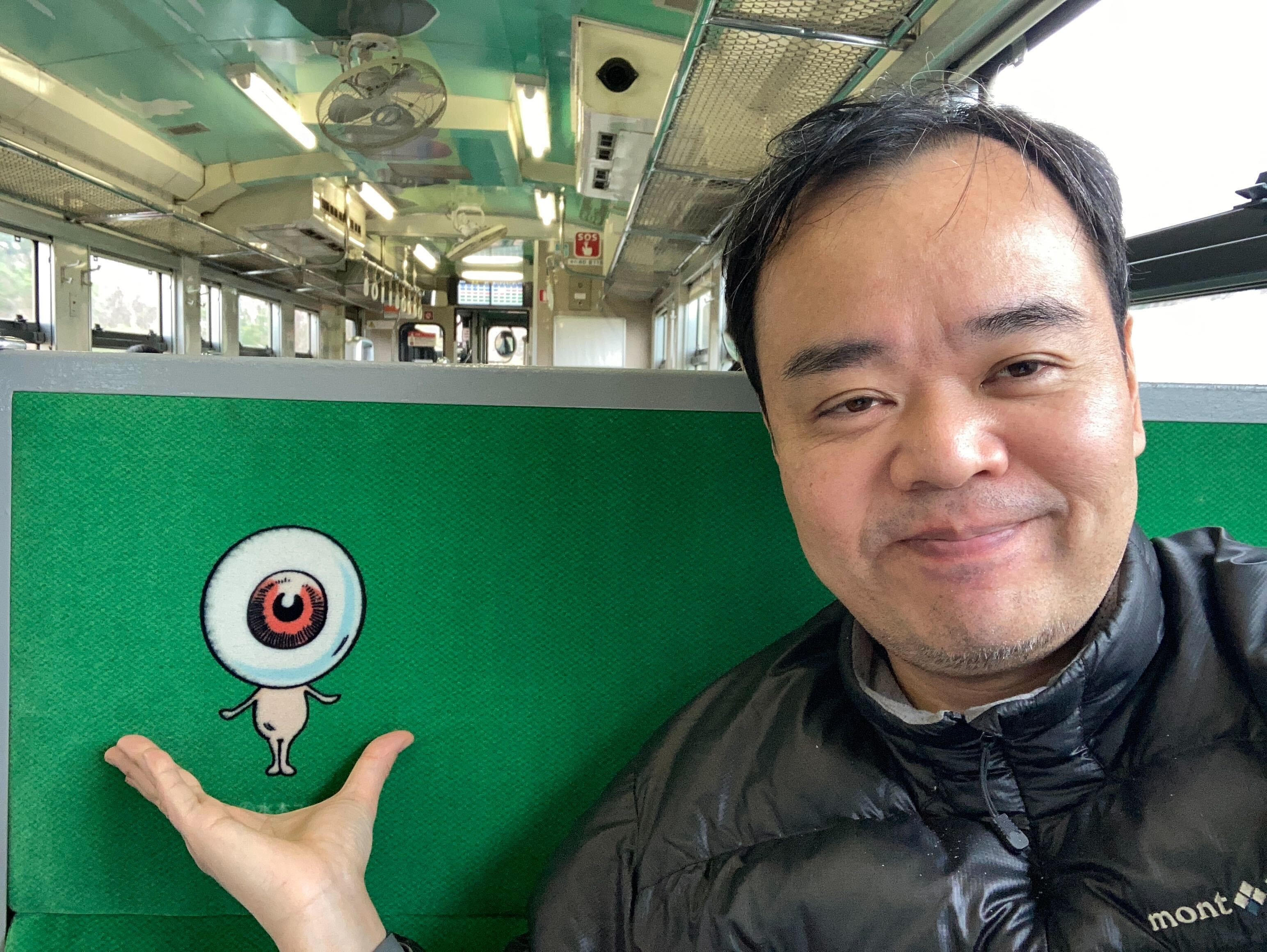 A man smiling and sitting on a train next to a cartoon eyeball character illustration on a green seat back.