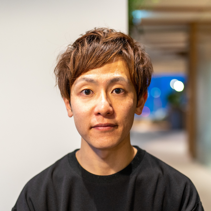 A portrait of an Asian man with short hair, wearing a black shirt, photographed indoors with a blurred background.