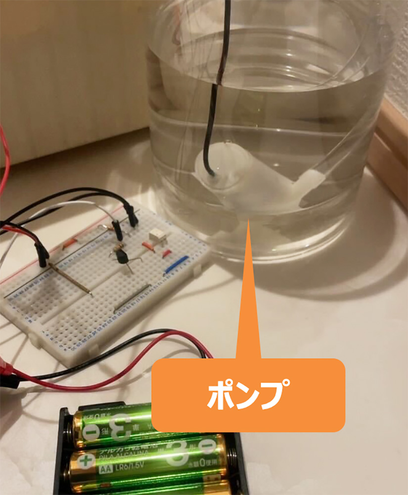 A smart gardening prototype setup showing a transparent container with a submersible water pump, simple breadboard circuit, and battery pack. The Japanese label ポンプ highlights the pump used in the system.