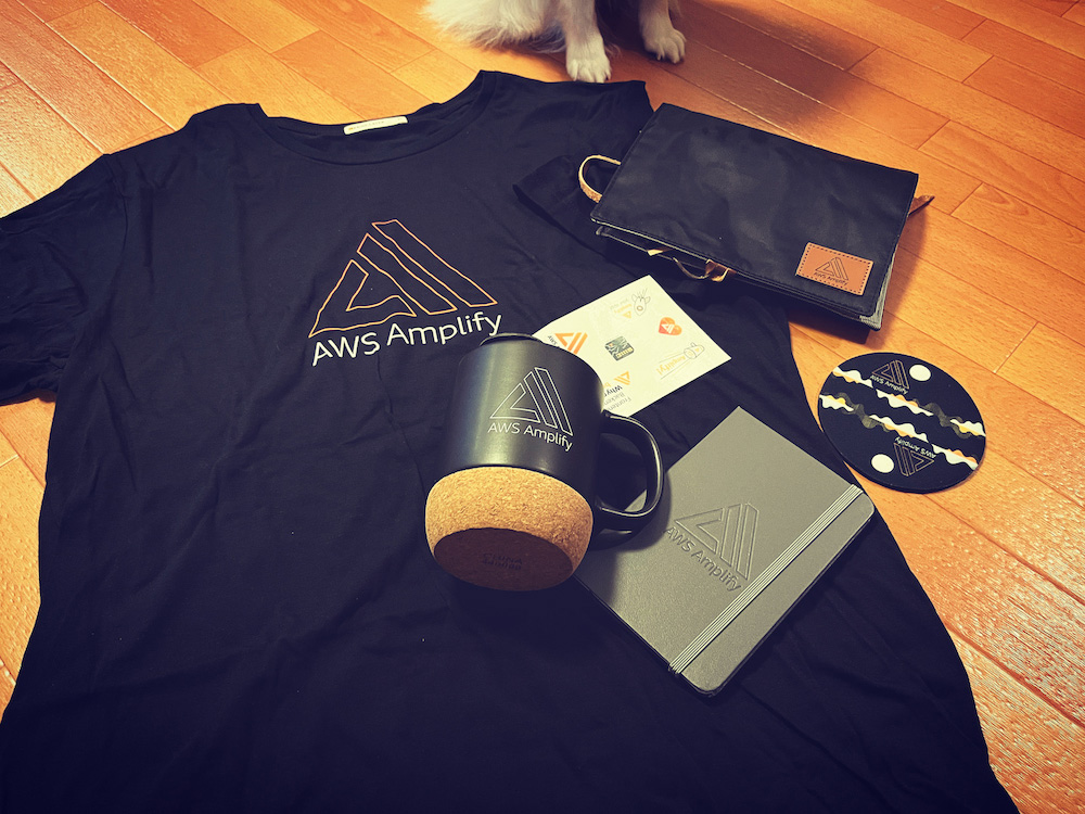 A collection of AWS Amplify branded swag items including a black t-shirt, mug, notebook, stickers, a pouch, and coasters displayed on a wooden floor.