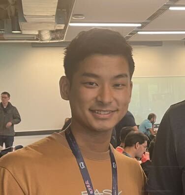 A young man wearing a brown shirt and lanyard is smiling at the camera in an indoor event setting, with people and desks in the background.