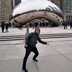 A person wearing a hooded jacket poses playfully in front of Cloud Gate (The Bean) in Chicago, with people and city buildings in the background.