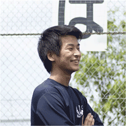 A man smiling outdoors, standing in front of a fence with greenery and a Japanese sign in the background.