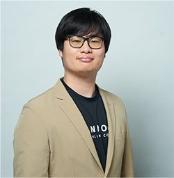 Portrait of a man with black hair wearing glasses and a beige blazer over a black T-shirt, standing against a light background.