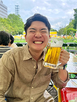 A person smiling broadly while holding a glass of beer at an outdoor festival, seated at a table with people and greenery in the background.