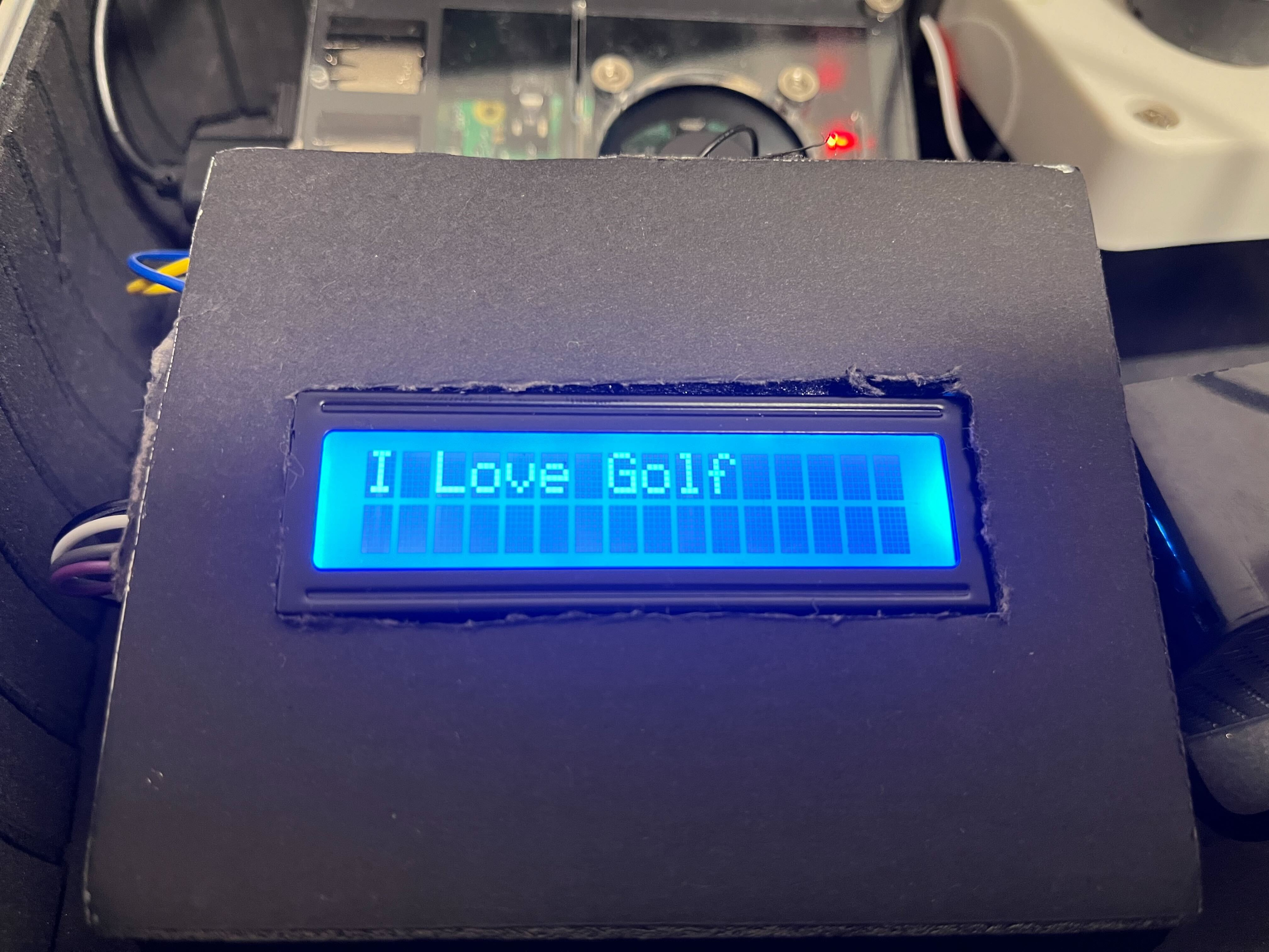 A close-up image of an electronic LCD display showing the message 'I Love Golf' in blue text, mounted on a dark panel with visible wiring and electronic components in the background.