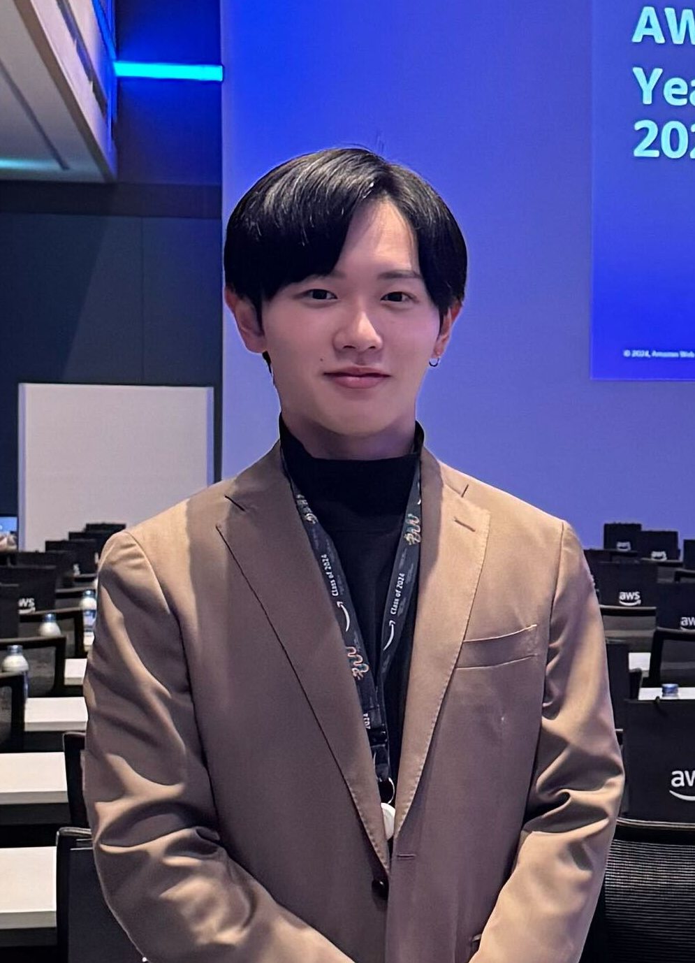 A person wearing a brown blazer and a lanyard stands in an AWS event venue with conference tables and AWS branding visible in the background.
