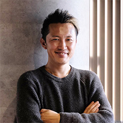 A casual portrait of a man standing with arms crossed, smiling, in front of a textured wall and window blinds.