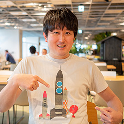 A person in a casual office setting wearing a white t-shirt with colorful rocket illustrations, pointing at the shirt and smiling.