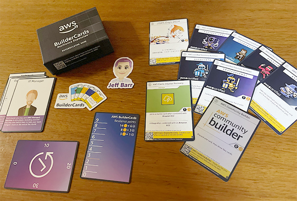 A photo of the AWS BuilderCards Japanese Edition card game, featuring a black box labeled 'AWS BuilderCards,' various playing cards, a character cutout, and game pieces spread out on a wooden surface.