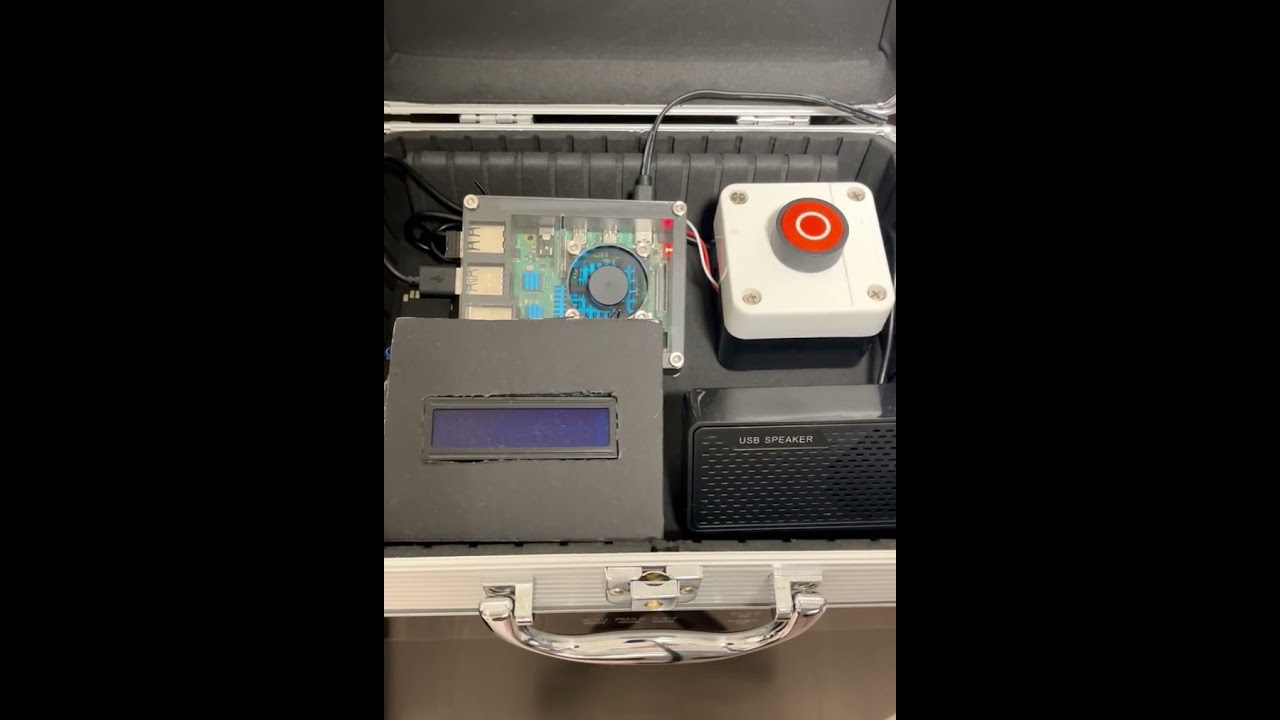 An open electronics case showing a DIY setup including a circuit board, a large red button, an LCD screen, and a USB speaker.