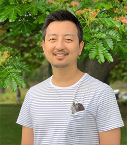 A man standing outdoors in front of a tree, wearing a white and gray striped t-shirt, smiling at the camera.
