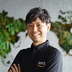 Professional portrait of an AWS employee wearing a branded AWS shirt, smiling, with a green plant wall in the background.