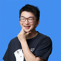A smiling man wearing glasses and a dark t-shirt with a cartoon graphic, posing with his hand on his chin in front of a blue background.