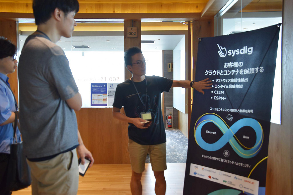 A presenter explains Sysdig cloud and container security solutions to attendees at JAWS DAYS 2023. The group is gathered around a promotional banner describing Sysdig's cloud and container protection features.