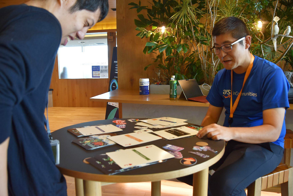Two participants engage in a card game discussion at an AWS Communities event, sitting at a table covered with cards and materials in a modern, plant-filled indoor setting.