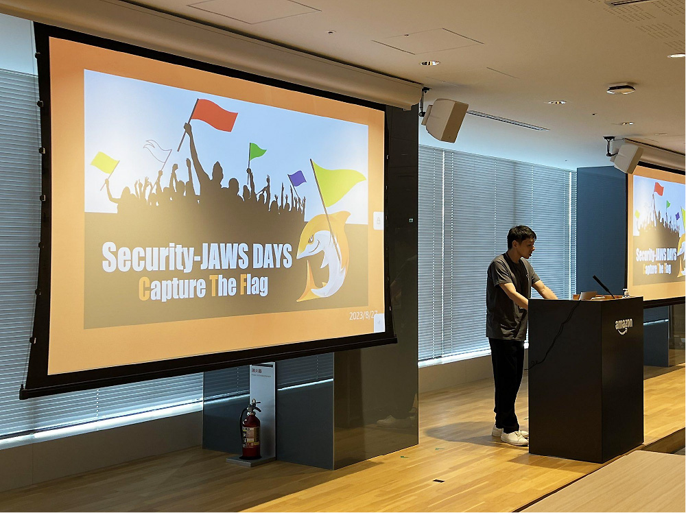 A presenter stands at a podium in an Amazon office, giving a presentation during the Security-JAWS DAYS Capture the Flag event. The projected screen displays the event title with an image of cheering silhouettes holding flags and a stylized shark logo.