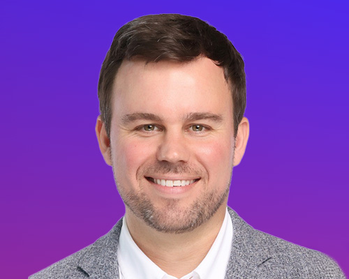 A professional headshot of a smiling man with dark hair and a trimmed beard, wearing a light gray blazer and white shirt, set against a purple gradient background.