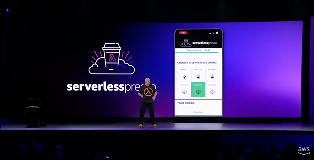 A presenter stands on stage in front of a large screen displaying the AWS Serverlesspresso logo and a mobile demo app interface. The presentation showcases serverless drink order selection as part of an AWS event demo.