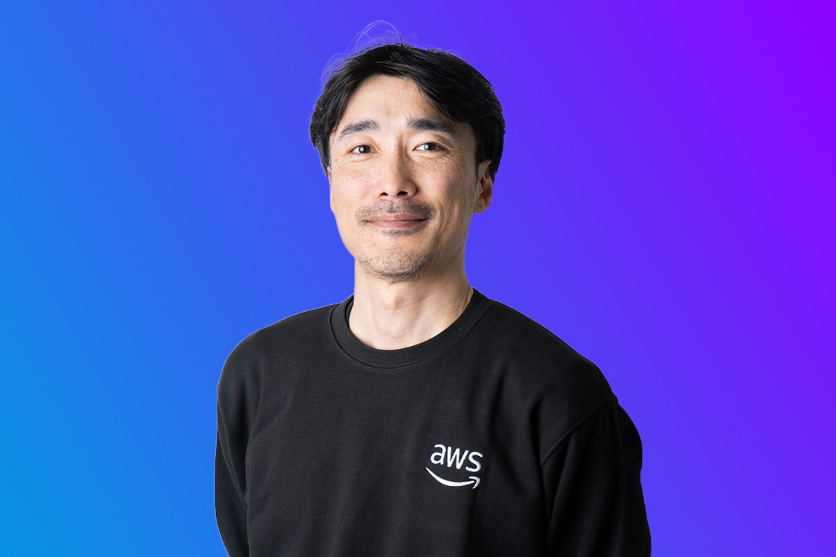 A portrait of an AWS presenter wearing a black AWS logo sweatshirt, positioned in front of a blue-to-purple gradient background, for use in Japan Summit 2025 event promotions.
