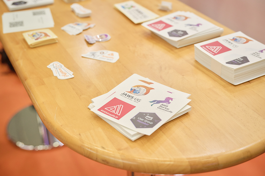 A variety of AWS User Group and community-themed stickers are displayed on a wooden table, likely at an AWS Village or community event.