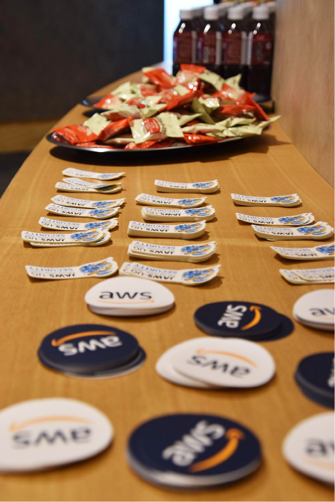 Table display featuring AWS security event badges, branded stickers, snacks, and bottled drinks, prepared for event participants.