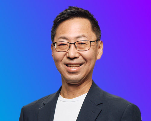 Portrait of a male speaker in a dark blazer and glasses against a blue to purple gradient background, AWS Summit Japan 2025.