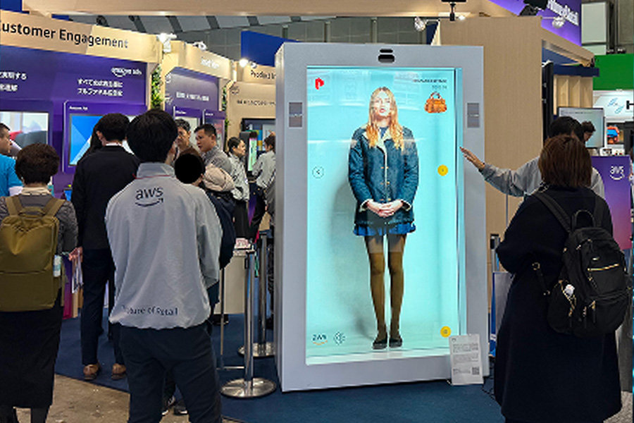 Attendees interact with a large digital display featuring an avatar at the AWS Summit Tokyo 2025 expo hall, with various exhibits and customer engagement booths in the background.