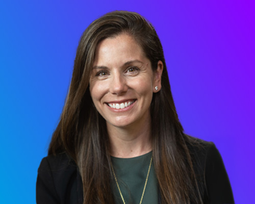 A professional woman with long brown hair, wearing a dark blazer and a green top with a necklace, smiling against a blue and purple gradient background.