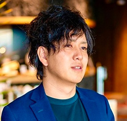 A portrait of a person with medium-length dark hair, wearing a blue casual blazer over a green shirt, photographed indoors with a softly blurred background.