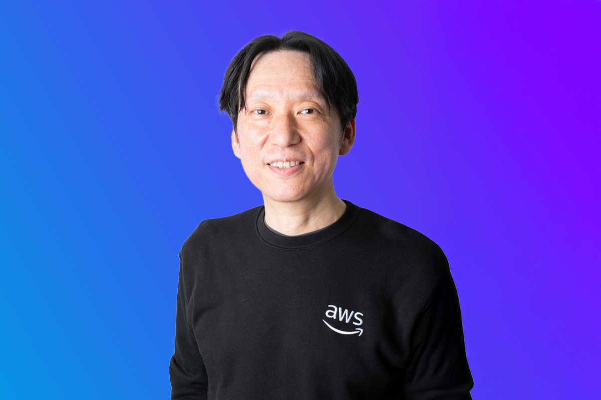 A person wearing an AWS sweater, smiling in front of a blue to purple gradient background. Used for AWS Japan Summit 2025 event promotional materials.