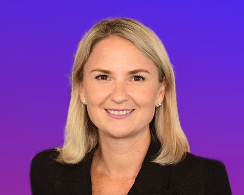 A professional woman with blonde hair, wearing a black blazer, is smiling in front of a purple gradient background.