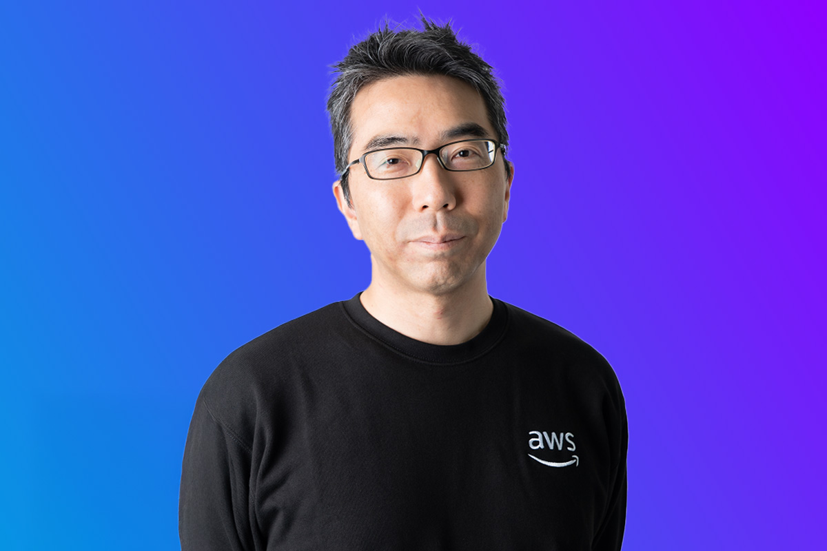 A man wearing glasses and an AWS-branded black shirt stands in front of a blue to purple gradient background. This image appears to be a portrait, possibly for a speaker or attendee at an AWS Japan Summit event.