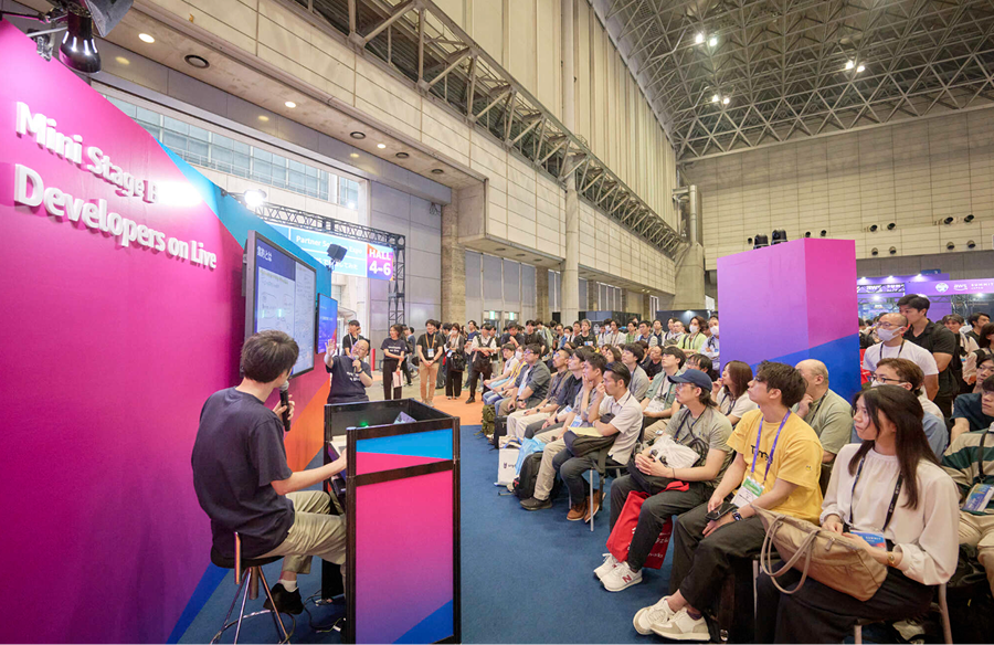 A presenter speaks to a large audience at the Mini Stage Developers on Live event during AWS Summit 2025, held in a spacious expo hall with attendees seated and watching a technical demonstration or talk.