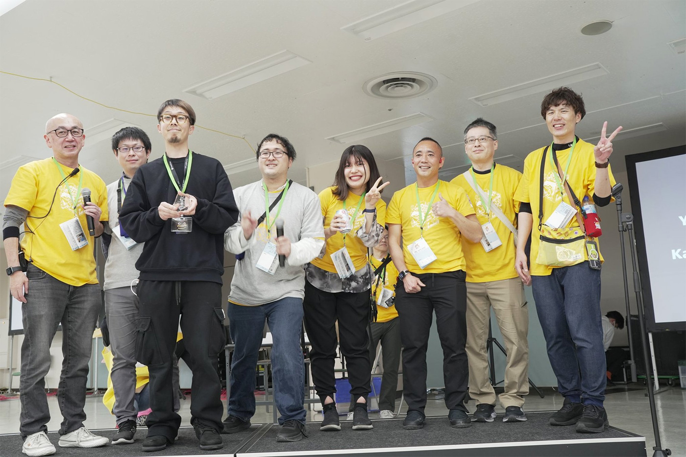A group of participants and presenters pose for a group photo on stage during the AWS Samurai 2024 event. Some individuals are wearing yellow event shirts, holding microphones and event materials, and smiling towards the camera.