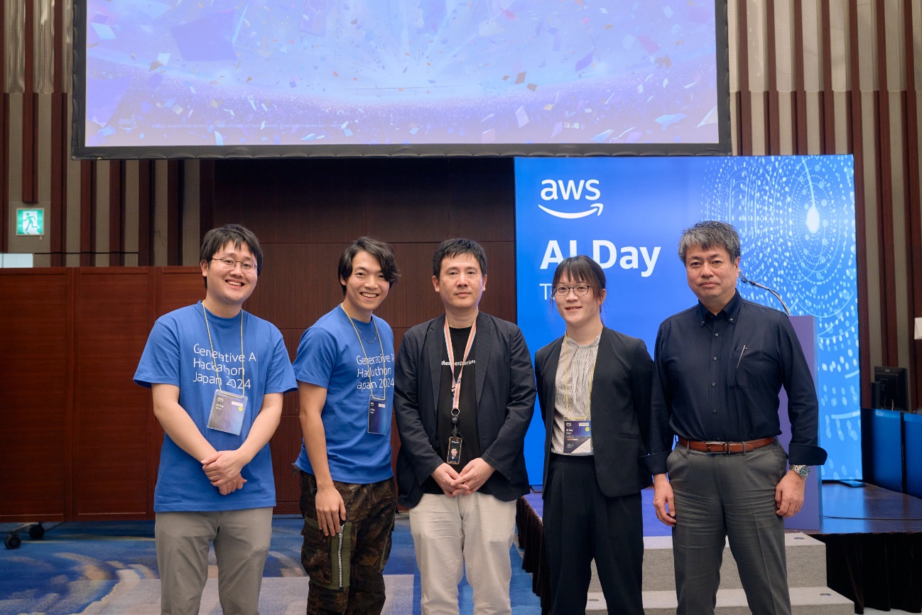 Group photo of participants at AWS AI Day Generative AI Hackathon Japan 2024, featuring several individuals standing in front of an event backdrop with 'AWS AI Day' signage.