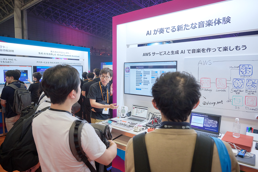 People gather around an AWS booth at an expo in Japan, where a presenter demonstrates how to use AWS services and generative AI to create music. The booth features monitors displaying a song generator interface, diagrams, and technical equipment, promoting an innovative AI-powered music experience.
