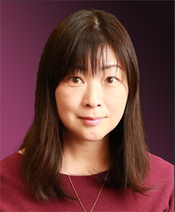Professional headshot of a woman with straight brown hair, wearing a burgundy top, in front of a purple gradient background.