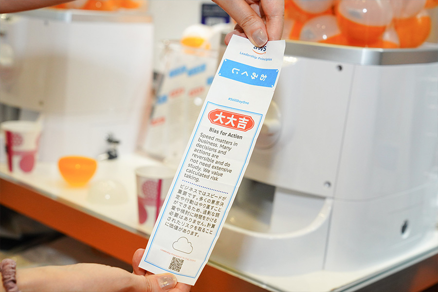 A close-up of a person holding an Omikuji slip at the AWS Summit 2025 OneAWS Zone. The slip features 'Bias for Action' leadership principle in English and Japanese, with colorful capsules, cups, and a machine in the background.